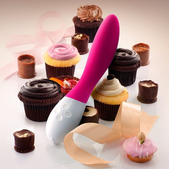 Vibromasseur point G Mona 2 rose pose parmi des cupcakes et rubans, mise en scene gourmande du masseur Lelo en silicone a la poignee blanche ergonomique.