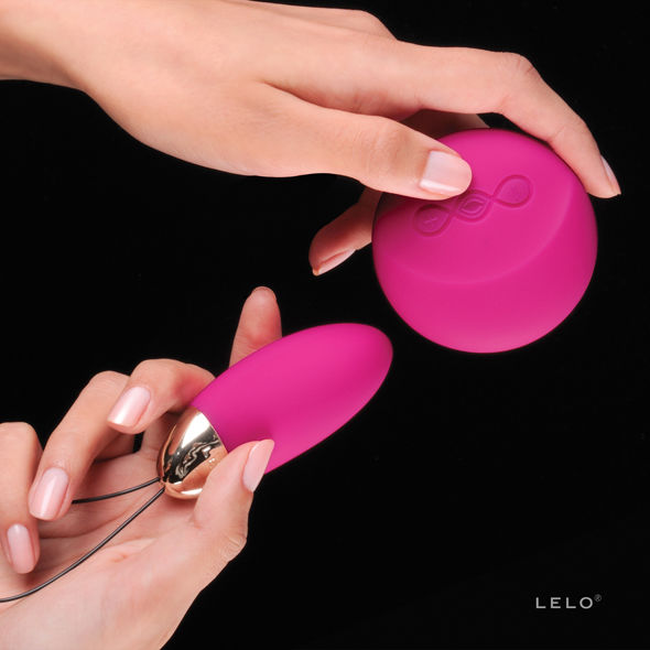 Mains tenant l'œuf vibrant Lyla 2 de Lelo rose et sa télécommande ronde dorée, gros plan sur le silicone lisse et le cordon de retrait sur fond noir.