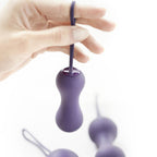 Main retenant une boule de Kegel violette double par son cordon de retrait, autres billes floues en arrière-plan, démonstration d'usage du produit sur fond blanc parfaitement épuré.