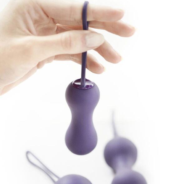 Main retenant une boule de Kegel violette double par son cordon de retrait, autres billes floues en arrière-plan, démonstration d'usage du produit sur fond blanc parfaitement épuré.